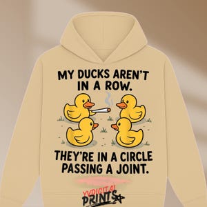 May include: A beige hooded sweatshirt featuring a graphic of four yellow rubber ducks in a circle. The text reads: MY DUCKS AREN'T IN A ROW. THEY'RE IN A CIRCLE PASSING A JOINT. One duck holds a lit joint with smoke rising above it. YVDIGITAL PRINTS is at the bottom.