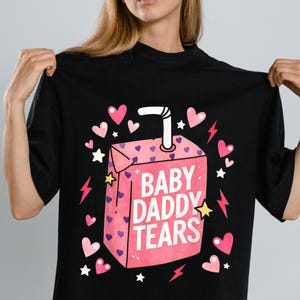 May include: Black t-shirt featuring a pink juice box graphic with the text "BABY DADDY TEARS." The juice box is decorated with pink hearts, white stars, and pink lightning bolts. A white straw extends from the top of the juice box.