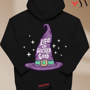 May include: Black hoodie with a purple witch's hat graphic and the words "Life is Wicked Good" in white. The hat has a green band and buckle. White star accents surround the hat, creating a festive design.