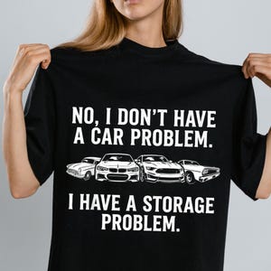 May include: Black t-shirt with the text "NO, I DON'T HAVE A CAR PROBLEM. I HAVE A STORAGE PROBLEM." The shirt features illustrations of four different cars in white. A humorous design for car enthusiasts.