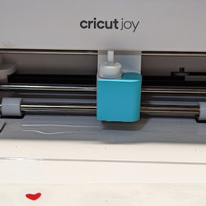 May include: A white Cricut Joy machine with a teal cutting head. The machine has silver rollers and the brand name is printed in black. The machine is designed for crafting and DIY projects.