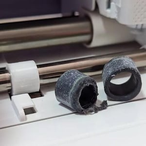 May include: A close-up shot of two worn, dark grey rubber rollers with a cylindrical shape, next to a white plastic component and a silver metal rod. The rollers appear damaged, with one showing signs of wear and tear.
