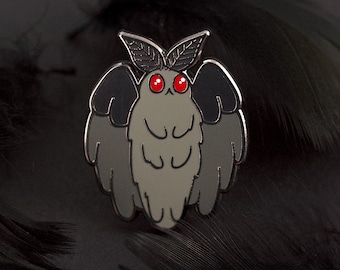 May include: A black and gray enamel pin of a moth with red eyes. The moth is on a black background with black feathers.