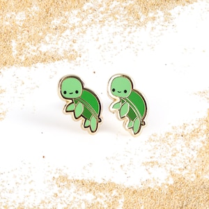 May include: A pair of gold-tone turtle earrings with green enamel. The turtles have smiling faces and are facing each other.