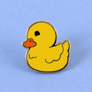 May include: A yellow rubber duck pin with a black eye and orange beak. The duck is sitting on a light blue background.