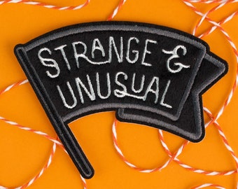May include: A black and gray embroidered patch with the words "STRANGE & UNUSUAL" in white script. The patch is in the shape of a flag and is set against an orange background with white and orange twine.