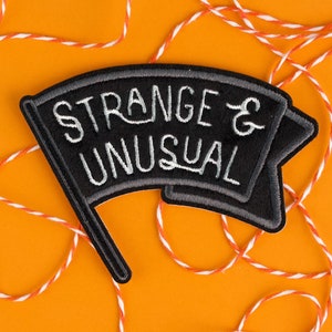 May include: A black and gray embroidered patch with the words "STRANGE & UNUSUAL" in white script. The patch is in the shape of a flag and is set against an orange background with white and orange twine.