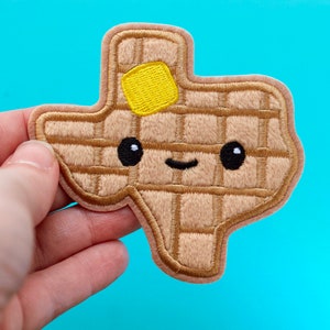 May include: A tan embroidered patch in the shape of the state of Texas, resembling a waffle. It features a yellow butter square and a smiling face with black eyes. The patch is held against a blue background.
