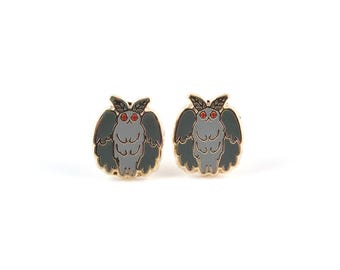 May include: A pair of moth-shaped enamel pins. Each pin features a gray moth with dark green wings, red eyes, and a gold-colored border. The pins are set against a white background.