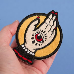 May include: A yellow and black embroidered patch with a white hand with a red eye in the palm. The hand is in a praying position.