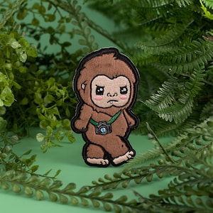 May include: Brown felt patch of a cartoon Bigfoot with a green strap and a camera around its neck. The Bigfoot has a grumpy expression and is looking to the right.