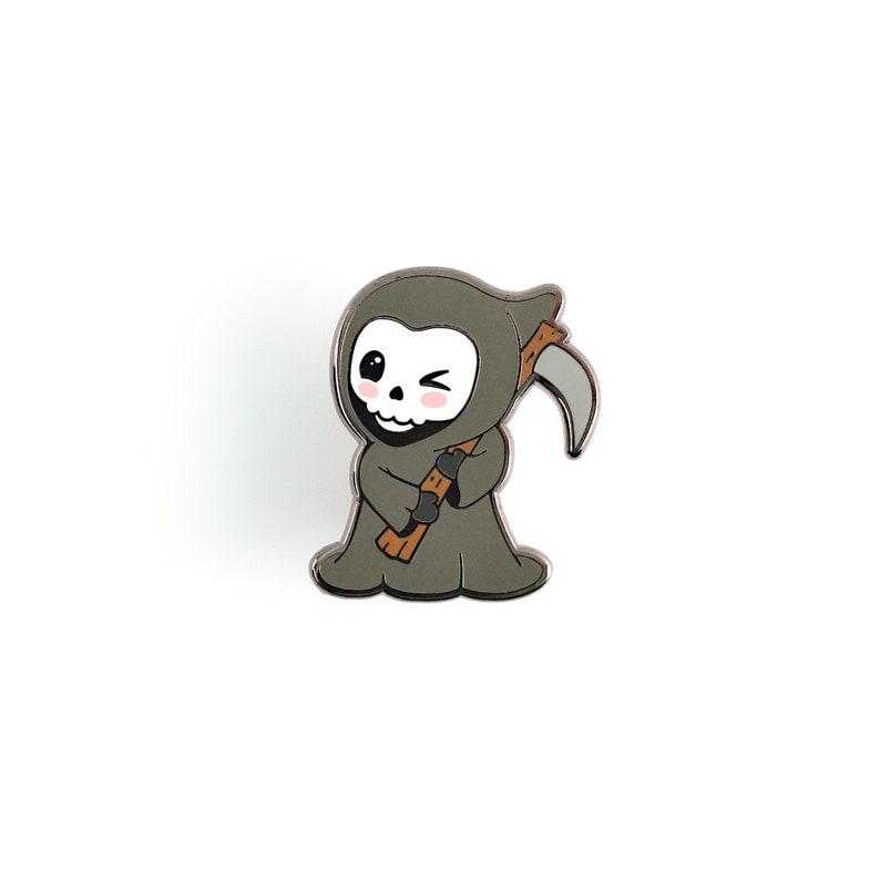 May include: A cute enamel pin featuring a cartoon Grim Reaper character. The pin has a gray robe, a scythe, and a winking face with rosy cheeks. The pin is set against a white background.