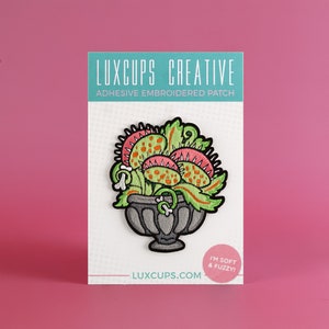 May include: A green, pink, and orange embroidered patch of a Venus flytrap plant in a gray pot. The patch is on a white card with the text "LUXCUPS CREATIVE ADHESIVE EMBROIDERED PATCH LUXCUPS.COM I'M SOFT & FUZZY!"