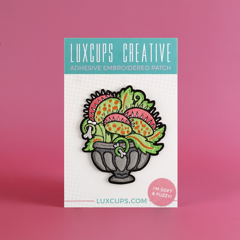 May include: A green, pink, and orange embroidered patch of a Venus flytrap plant in a gray pot. The patch is on a white card with the text "LUXCUPS CREATIVE ADHESIVE EMBROIDERED PATCH LUXCUPS.COM I'M SOFT & FUZZY!"