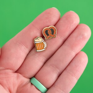 May include: Two gold enamel pins, one depicting a pretzel and the other a beer mug. The pretzel is brown and the beer mug is orange.