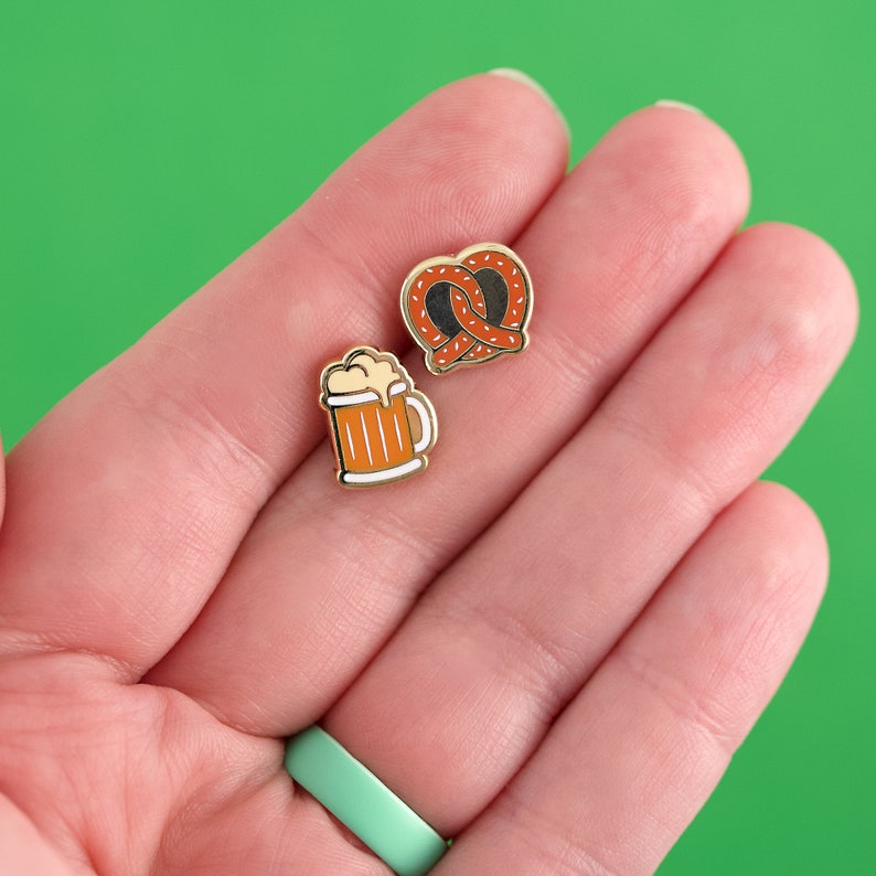 May include: Two gold enamel pins, one depicting a pretzel and the other a beer mug. The pretzel is brown and the beer mug is orange.