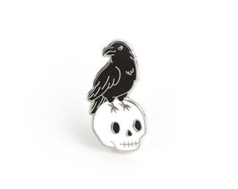 May include: Black and white enamel pin featuring a black raven perched on a white skull.