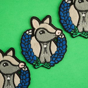 May include: Embroidered patches featuring a cartoon armadillo wearing a star-shaped bolo tie and a blue wreath. The patches have a gray, tan, and blue color scheme, with a green background.