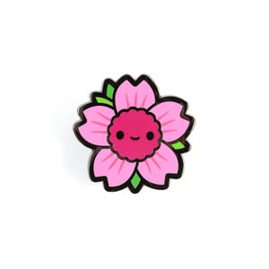 May include: A pink flower-shaped enamel pin with a smiling face in the center. The pin has a black outline and green accents on the petals. The flower design is a cute and cheerful accessory.