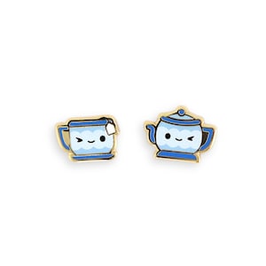 May include: A pair of gold-toned earrings featuring a blue teacup and teapot design. The teacup has a tea bag and the teapot has a lid. Both have smiling faces and are outlined in gold.