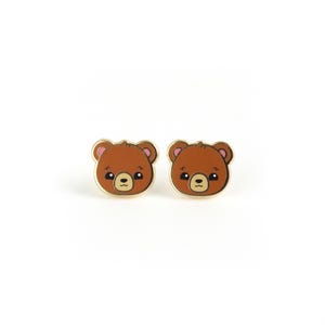 May include: A pair of gold-toned stud earrings featuring a cartoon bear face design. The bear has brown fur, pink cheeks, and black eyes.