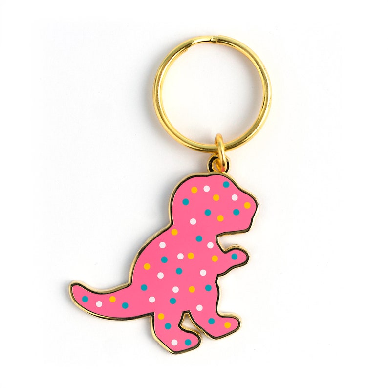 May include: A pink dinosaur-shaped keyring with colourful sprinkles. The keyring has a gold metal ring.
