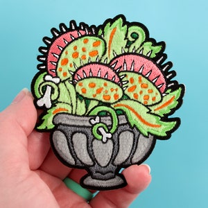 May include: A green, pink, and orange embroidered patch of a carnivorous plant in a gray pot. The plant has sharp teeth and is in a vintage style.