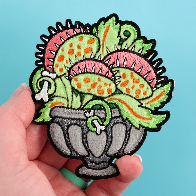 May include: A green, pink, and orange embroidered patch of a carnivorous plant in a gray pot. The plant has sharp teeth and is in a vintage style.