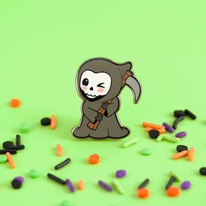 May include: A gray enamel pin of a winking cartoon Grim Reaper holding a scythe. The pin has a black outline and a light pink blush on the cheeks. The background is a bright green with colorful sprinkles.