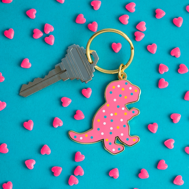 May include: A pink and white dinosaur keyring with gold accents. The keyring is attached to a silver key. The keyring is surrounded by pink hearts on a blue background.