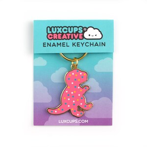 May include: Pink dinosaur shaped enamel keychain with colourful sprinkles. The keychain is attached to a gold metal ring. The keychain is packaged on a card with the text "LUXCUPS CREATIVE ENAMEL KEYCHAIN LUXCUPS.COM".