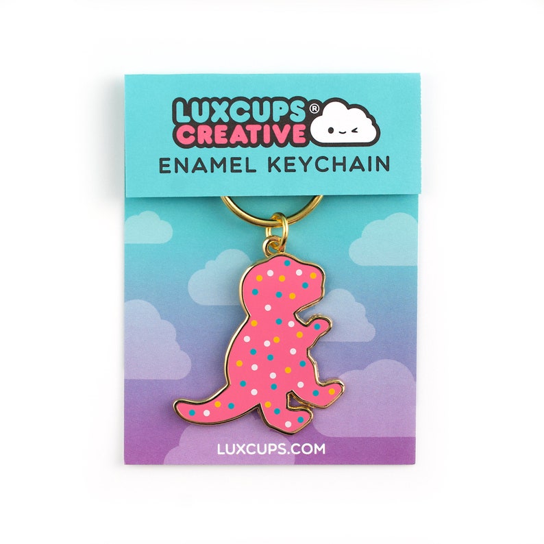 May include: Pink dinosaur shaped enamel keychain with colourful sprinkles. The keychain is attached to a gold metal ring. The keychain is packaged on a card with the text "LUXCUPS CREATIVE ENAMEL KEYCHAIN LUXCUPS.COM".