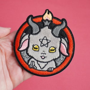 May include: A gray embroidered patch of a cute cartoon goat with horns, a pentagram on its forehead, and a red circle around it.