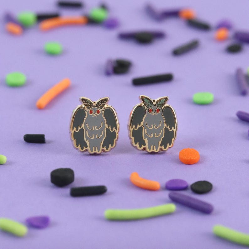 K&ouml;nnte beinhalten: Zwei Ohrstecker in Mottenform mit goldenen R&auml;ndern, grauen K&ouml;rpern und schwarzen Fl&uuml;geln, vor einem lila Hintergrund mit bunten Streuseln. Die Ohrringe haben rote Augen und sind ein einzigartiges Accessoire.