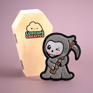 May include: A gray embroidered patch of a cartoon Grim Reaper with a scythe, winking and smiling. The patch is on a wooden coffin with the text "LUXCUPS CREATIVE" printed on it.