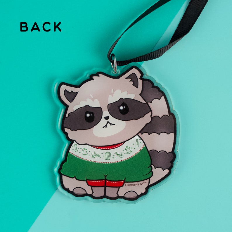 Puede incluir: Un adorno navide&ntilde;o con un mapache de dibujos animados que lleva un su&eacute;ter verde con un dise&ntilde;o festivo. El mapache tiene una expresi&oacute;n gru&ntilde;ona y lleva pantalones rojos. El adorno est&aacute; hecho de acr&iacute;lico transparente y tiene una cinta negra atada a la parte superior.