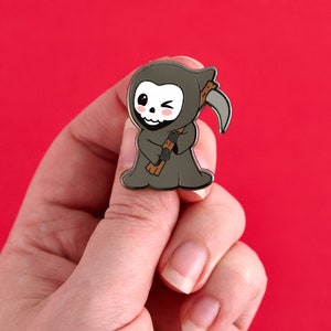May include: A cute enamel pin featuring a cartoon Grim Reaper character. The reaper is gray with a winking skull face, holding a scythe and wearing a hooded cloak. The pin is held against a red background.