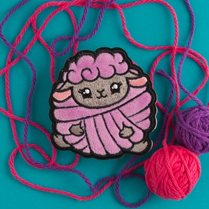 May include: A pink and gray embroidered sheep patch with big eyes. The patch is surrounded by pink and purple yarn.