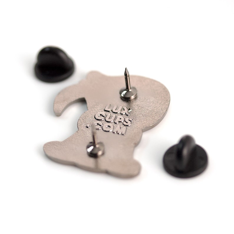 May include: A silver-colored enamel pin with a cartoon design, featuring a pin back and two black pin clasps. The text "LUXCUPS.COM" is stamped on the back of the pin. The pin is on a white background.