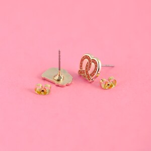 May include: A pair of gold-tone earrings with a pretzel design. The earrings are on a pink background.
