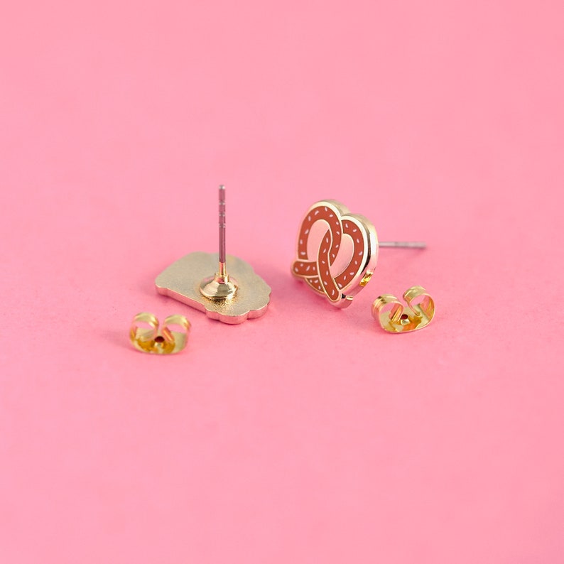 May include: A pair of gold-tone earrings with a pretzel design. The earrings are on a pink background.