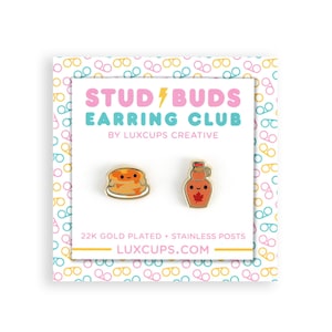 May include: A pair of gold-plated earrings featuring a cartoon pancake and a bottle of maple syrup. The earrings are part of the Stud/Buds Earring Club by Luxcups Creative. The text "22K Gold Plated - Stainless Posts" and "LUXCUPS.COM" are printed on the card.