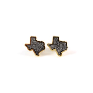 May include: A pair of gold-toned stud earrings shaped like the state of Texas. The earrings are filled with black glitter.
