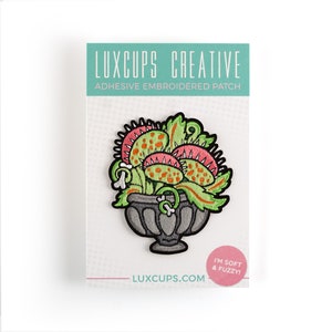 May include: A green, pink, and orange embroidered patch of a Venus flytrap plant in a gray pot. The patch is labeled "Luxcups Creative Adhesive Embroidered Patch" and "I'm soft & fuzzy!"