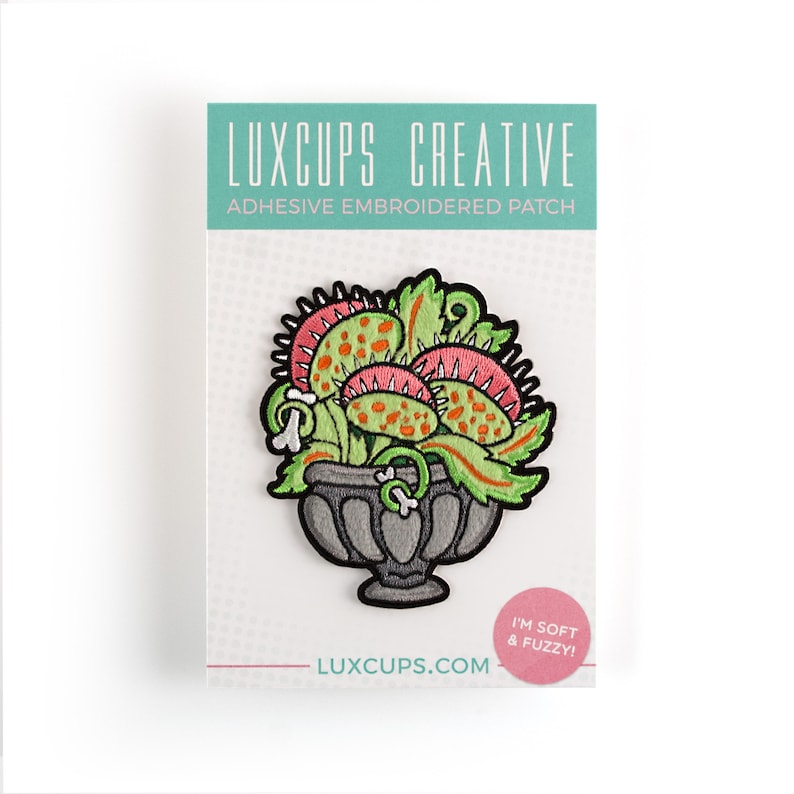 May include: A green, pink, and orange embroidered patch of a Venus flytrap plant in a gray pot. The patch is labeled "Luxcups Creative Adhesive Embroidered Patch" and "I'm soft & fuzzy!"