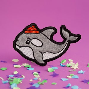 May include: A gray and white embroidered patch of a cartoon orca wearing a red and orange hat. The patch is on a purple background with confetti.