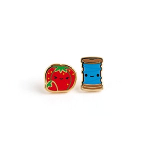 May include: Two gold-colored enamel pins, one shaped like a strawberry with a red and green design and a smiling face, the other shaped like a spool of thread with a blue thread and a smiling face.