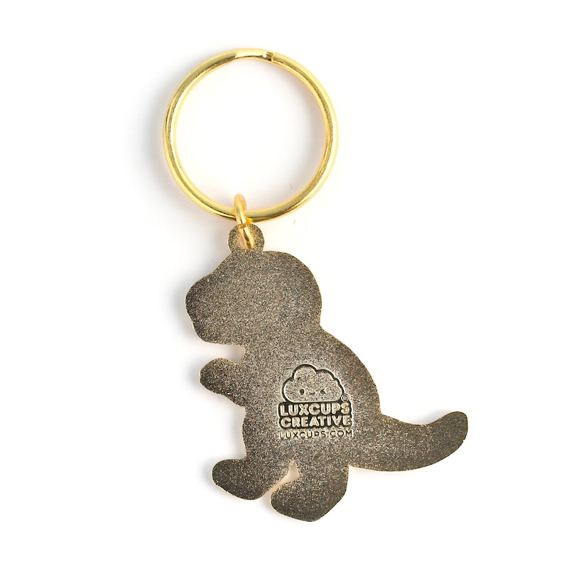 May include: Gold-coloured metal dinosaur keychain with a gold key ring. The back of the keychain has the text "LUXCUPS CREATIVE LUXCUPS.COM" printed on it.
