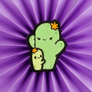 May include: A green felt cactus with a smaller green felt cactus attached to its side. Both cacti have smiling faces and yellow felt flowers.