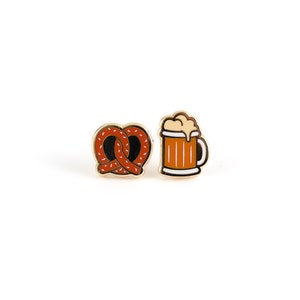 May include: A pair of gold-tone earrings featuring a pretzel and a beer mug. The pretzel is brown with white sprinkles and the beer mug is orange with a white foam top.